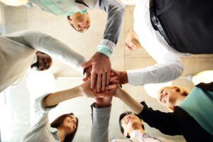 Group of adults standing in a circle with hands stacked together, smiling in a show of unity and support.