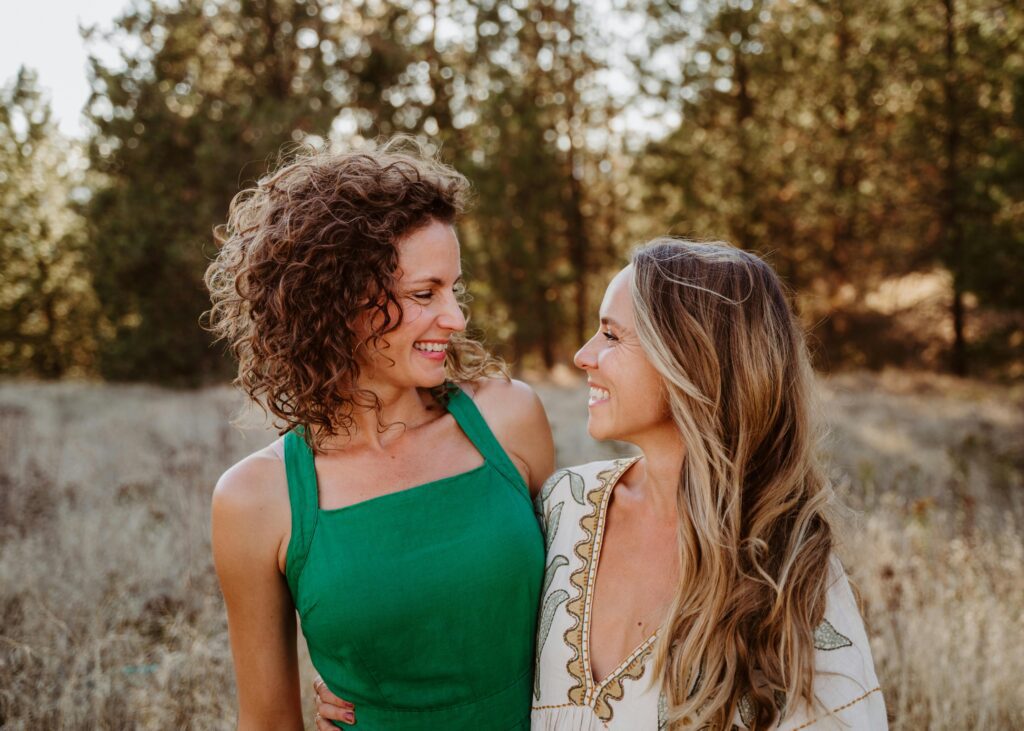 Two women smiling at each other outdoors in a sunlit natural setting.